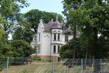 Historische weiße Villa mit spitzem Turm, umgeben von Bäumen und einem gepflegten Garten.