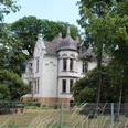 Historische weiße Villa mit spitzem Turm, umgeben von Bäumen und einem gepflegten Garten.