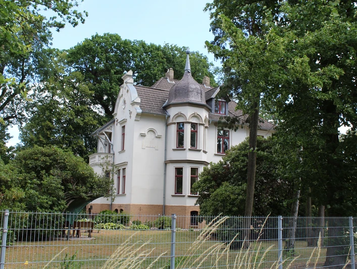 Historische weiße Villa mit spitzem Turm, umgeben von Bäumen und einem gepflegten Garten.