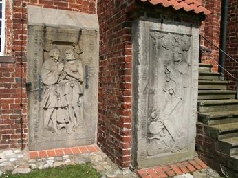 Reliefs aus Sandstein an einer roten Backsteinmauer mit Treppe, künstlerische Darstellungen von Personen.