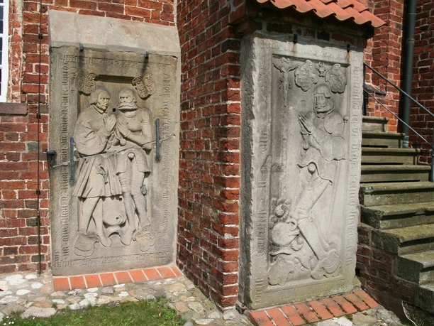 Reliefs aus Sandstein an einer roten Backsteinmauer mit Treppe, künstlerische Darstellungen von Personen.