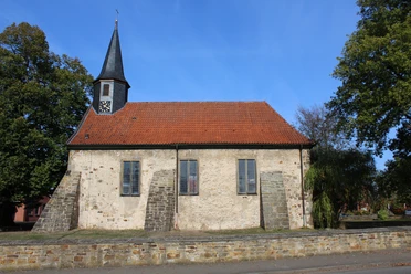 Historische Kirche Münchehagen mit rotem Ziegeldach und Uhrenturm, umgeben von Bäumen und Steinmauer.