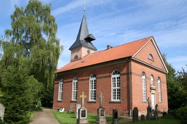 Backsteinkirche mit Spitzdach und Glockenturm, umgeben von Bäumen und Grabsteinen unter blauem Himmel.