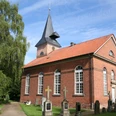 Backsteinkirche mit Spitzdach und Glockenturm, umgeben von Bäumen und Grabsteinen unter blauem Himmel.
