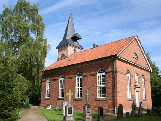 Backsteinkirche mit Spitzdach und Glockenturm, umgeben von Bäumen und Grabsteinen unter blauem Himmel.