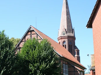 Ev.-luth. Kirche zu Estorf Ein Kirchturm mit Uhr ragt hinter grünen Bäumen und einem roten Backsteingebäude empor.