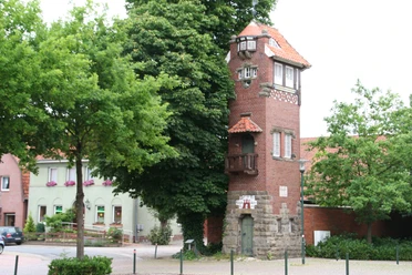 Feuerwehrturm Feuerwehrturm aus rotem Backstein mit weißen Fenstern und markantem Balkon, umgeben von Bäumen.