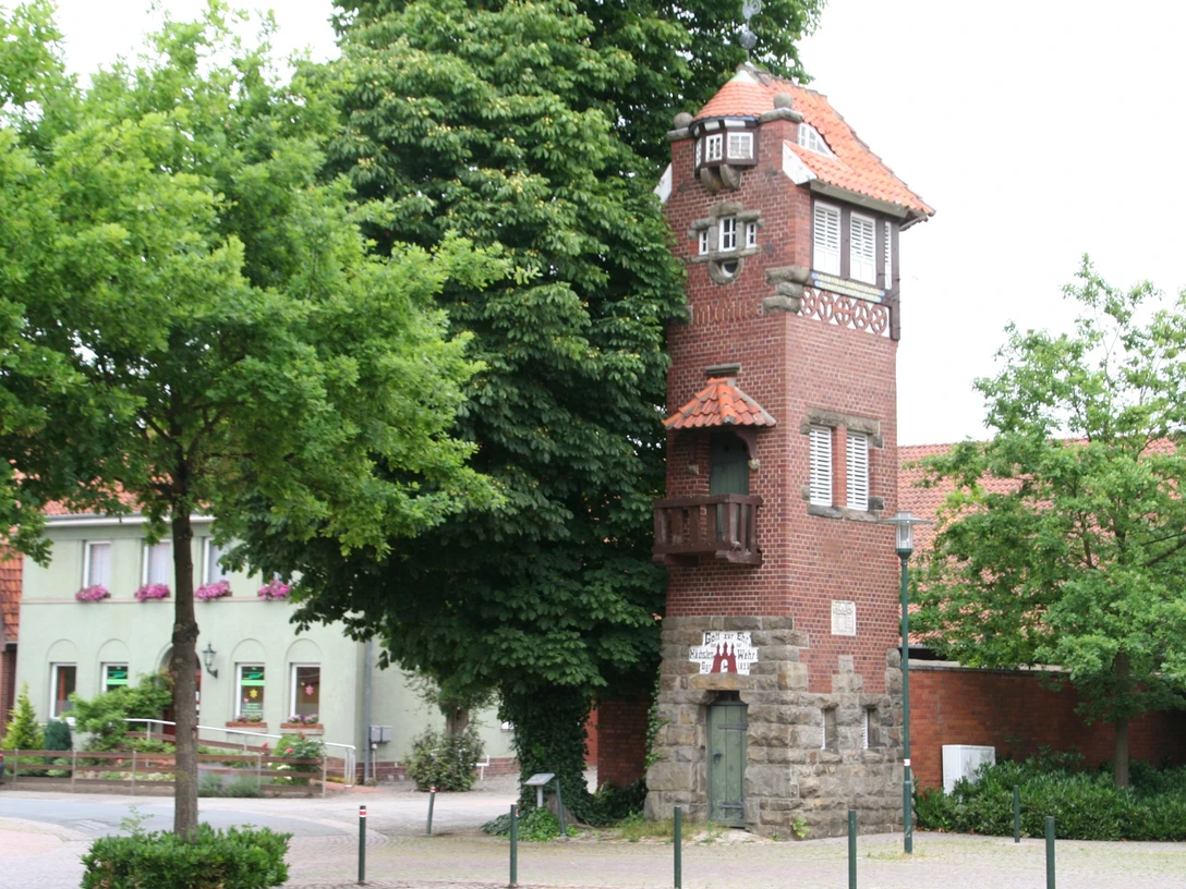 Feuerwehrturm Feuerwehrturm aus rotem Backstein mit weißen Fenstern und markantem Balkon, umgeben von Bäumen.