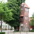 Feuerwehrturm Feuerwehrturm aus rotem Backstein mit weißen Fenstern und markantem Balkon, umgeben von Bäumen.