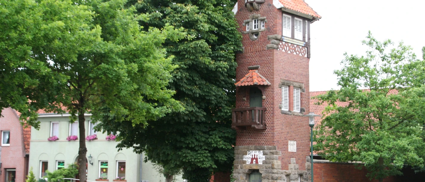 Feuerwehrturm Feuerwehrturm aus rotem Backstein mit weißen Fenstern und markantem Balkon, umgeben von Bäumen.