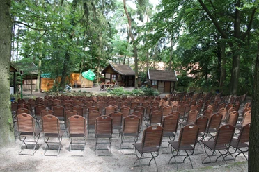 Freilichtbühne Stöckse Freilichtbühne mit leeren Holzbänken im Wald, umgeben von Bäumen und traditioneller Kulisse.