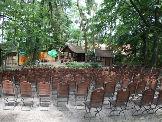 Freilichtbühne Stöckse Freilichtbühne mit leeren Holzbänken im Wald, umgeben von Bäumen und traditioneller Kulisse.