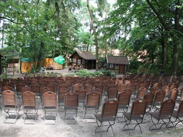 Freilichtbühne Stöckse Freilichtbühne mit leeren Holzbänken im Wald, umgeben von Bäumen und traditioneller Kulisse.