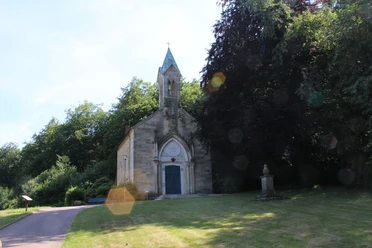 Eine historische Kapelle mit gotischen Elementen, umgeben von üppigem Grün und einem Gedenkstein.