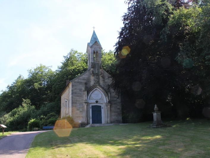 Friederikenkapelle Eine historische Kapelle mit gotischen Elementen, umgeben von üppigem Grün und einem Gedenkstein.
