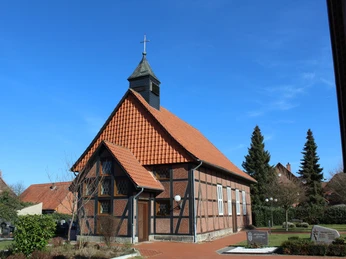 Kapelle Winzlar In einem Dorf steht eine kleine Fachwerkkapelle mit rotem Ziegeldach und begrünten, gepflegten Außenanlagen.