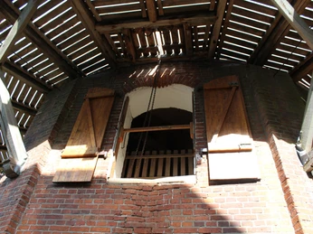 Roter Backsteinturm mit Holzbalkon, Fensterläden und offenen Holzlatten, Ketten sichtbar am Fenster.