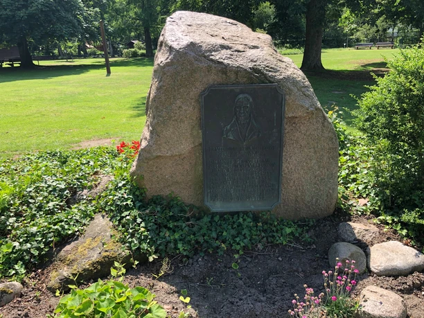 Gedenkstein mit Bronzetafel in einer grünen Parklandschaft, umgeben von kleinen Blumenbeeten.