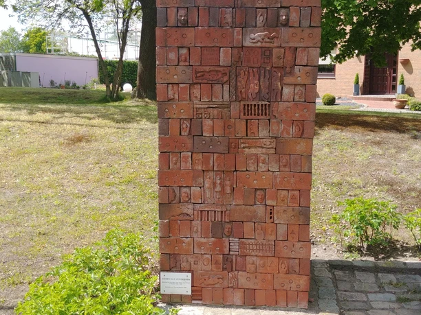 Gegen das Vergessen Ein hohes, rechteckiges Mosaikdenkmal aus roten Ziegeln mit eingravierten Symbolen und Figuren.