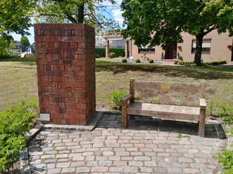 Gedenksäule aus roten Ziegeln neben einer hölzernen Bank auf gepflastertem Platz, umgeben von Bäumen.