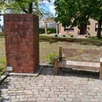 Gegen das Vergessen Gedenksäule aus roten Ziegeln neben einer hölzernen Bank auf gepflastertem Platz, umgeben von Bäumen.