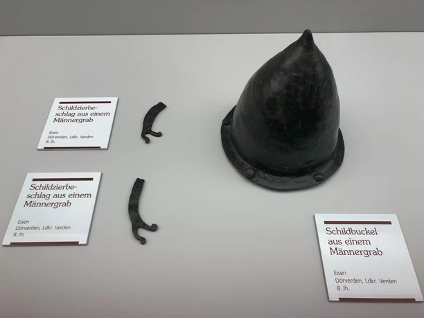 Schildbuckel und Schildzierbeschläge aus einem Männergrab - Ausstellungsstücke im Landesmuseum Hannover Ausstellungsstücke aus Eisen vom 8. Jh.: Schildbuckel und zwei Schildzierbeschläge, präsentiert im Landesmuseum Hannover.