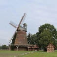 Historische Windmühle Hoyersförde vor bewölktem Himmel, umgeben von Bäumen und einem Fachwerkgebäude.