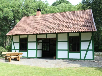 Fachwerkhaus mit grünem Gebälk und rotem Ziegeldach, umgeben von Bäumen und gepflastertem Weg.