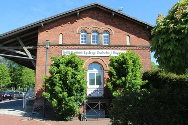 Güterschuppen Eystrup Der Güterschuppen Eystrup aus rotem Backstein mit grüner Bepflanzung vor zwei Rundbogenfenstern.