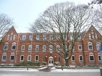 Altes rotes Backsteingebäude mit drei Giebeln und einem Baum im Vordergrund, umgeben von Schnee.