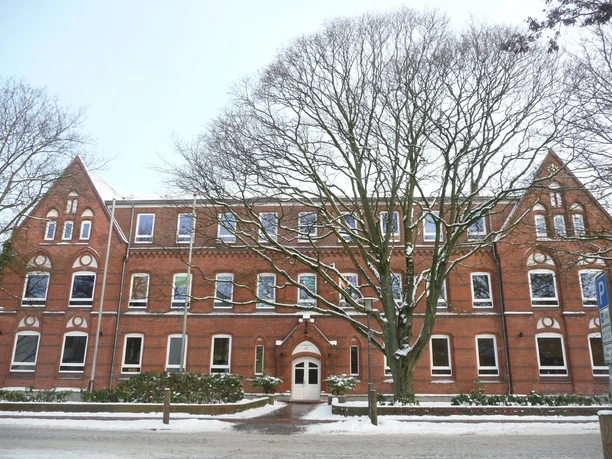 Altes rotes Backsteingebäude mit drei Giebeln und einem Baum im Vordergrund, umgeben von Schnee.