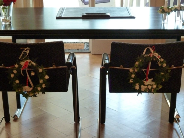 Zwei geschmückte Stühle stehen vor einem dunklen Tisch in einem lichtdurchfluteten Raum.