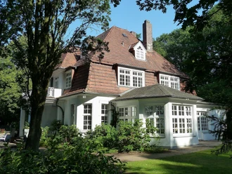 Ein elegantes historisches Gebäude mit großem Dach und hellen Fenstern, umgeben von üppigem Grün.