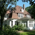 Haus Hünenburg Ein elegantes historisches Gebäude mit großem Dach und hellen Fenstern, umgeben von üppigem Grün.