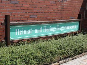 Heringsfängermuseum Schild Grünes Schild mit der Aufschrift "Heimat- und Heringsfängermuseum" vor roter Backsteinmauer.