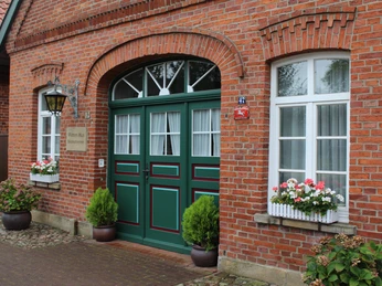 Rote Backsteinfassade mit grünen Türen, dekoriert mit Blumenkästen und Pflanzen zu beiden Seiten.