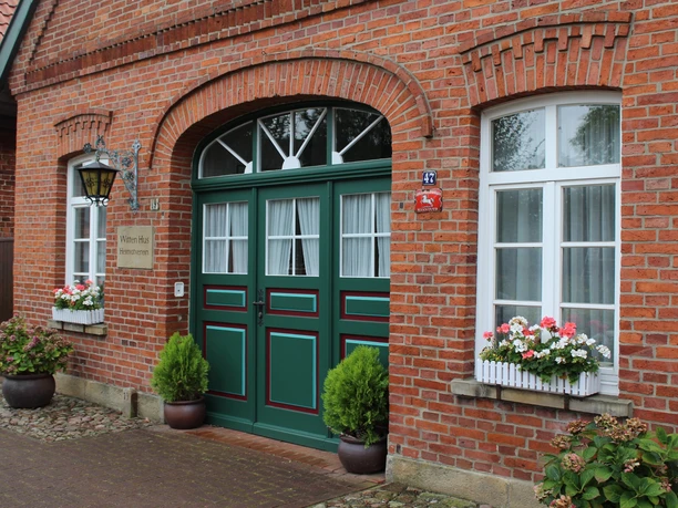 Rote Backsteinfassade mit grünen Türen, dekoriert mit Blumenkästen und Pflanzen zu beiden Seiten.