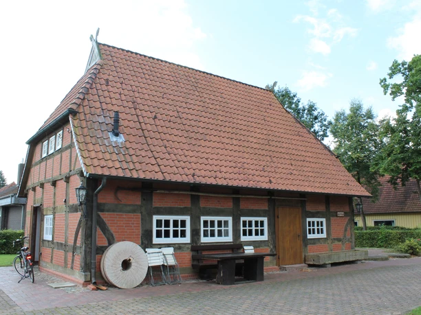 Fachwerkgebäude mit ziegelrotem Dach, umgeben von Bäumen und einem Fahrrad neben der Hauswand.