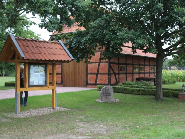 Historisches Fachwerkhaus mit rotem Ziegeldach, umgeben von Rasen, Bäumen und einem Informationsschild.