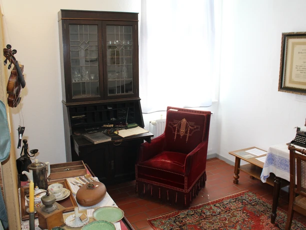 Ein gemütlicher Raum mit antiker Möblierung, darunter ein roter Sessel und eine Glasvitrine.