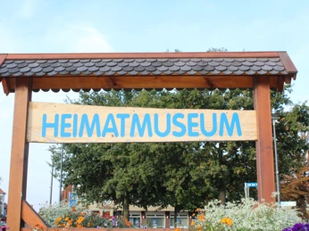 Holzschild mit blauer Aufschrift "Heimatmuseum" vor grünen Bäumen und bunten Blumen im Vordergrund.