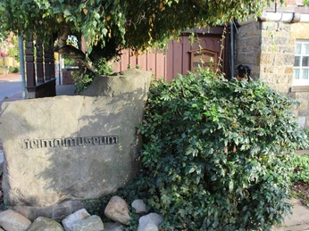 Heimatmuseum Rehburg Stein mit Inschrift "Heimatmuseum" vor Fachwerkgebäude umgeben von Sträuchern und Gehweg.