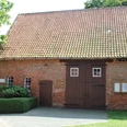 Ziegelsteinbau mit Satteldach in Wietzen, Eingang mit Doppeltür, umgeben von grüner Hecke.