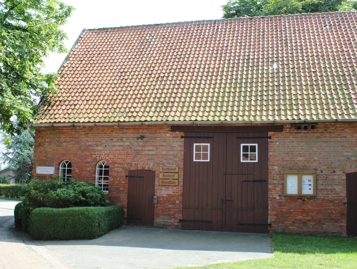 Ziegelsteinbau mit Satteldach in Wietzen, Eingang mit Doppeltür, umgeben von grüner Hecke.