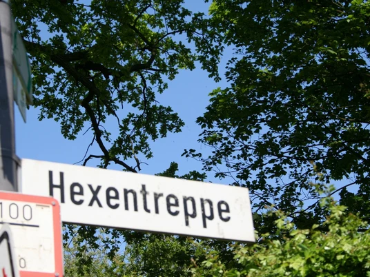 Ein Schild mit der Aufschrift "Hexentreppe", umgeben von üppigem Grün und blauem Himmel im Hintergrund.