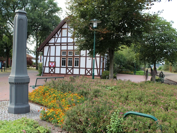 Historisches Fachwerkgebäude mit umliegendem Garten, Pflanzen und Parkbänken unter schattenspendenden Bäumen.