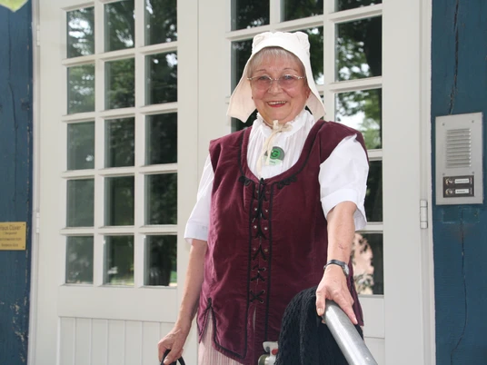 Ältere Frau in historischer Kleidung mit Lächeln, vor einer weiß gepflasterten Tür stehend.