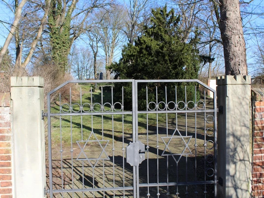 Metalltor mit Davidstern-Motiven öffnet den Zugang zu einem von Bäumen umgebenen Friedhof.