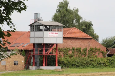 Historische Kartoffeldämpfanlage vor rotem Backsteingebäude mit grüner Kletterpflanze und Bäumen.