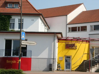 Kulturhaus mit rotem Dach und gelber Wand, Eingang über Treppe, Straßenschild und Fußgängerweg.